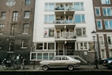 A vintage car is parked in front of an apartment building with multiple balconies. Bicycles are also lined up along the street, and a streetlamp is visible nearby. The buildings have a mix of brick and concrete finishes, giving the area a classic, urban feel.