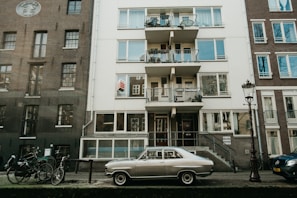 A vintage car is parked in front of an apartment building with multiple balconies. Bicycles are also lined up along the street, and a streetlamp is visible nearby. The buildings have a mix of brick and concrete finishes, giving the area a classic, urban feel.