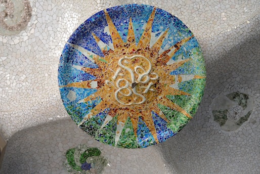 A colorful mosaic artwork featuring a circular design with a sun-like pattern at the center. The mosaic is composed of small, vibrant tiles in shades of blue, green, and gold, creating a radiant effect. The background is a contrasting, pale mosaic surface.