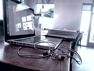 A laptop sits on a wooden table, accompanied by a pair of earphones resting on the surface. The background is slightly blurred, showing indistinct parts of the interior with large windows allowing soft light in. An open notebook lies next to the laptop, adding to the sense of a work or study environment.