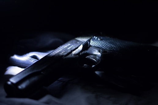 A handgun is partially illuminated against a dark background, with visible text on its surface. The lighting highlights its metallic texture, creating a dramatic and mysterious atmosphere.