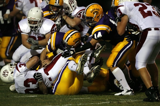 Players in a football game are engaged in a tackle, with athletes from opposing teams in action. The players are wearing helmets and protective gear, with some lying on the ground and others attempting to tackle or avoid the tackle. The setting appears to be an intense moment in the game.