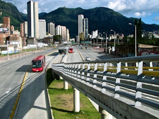 A bustling urban scene featuring a modern cityscape with tall buildings set against a backdrop of lush green mountains. A main roadway shows a dedicated bus lane with several red buses in transit, highlighting efficient public transportation. A pedestrian walkway is visible alongside the road.