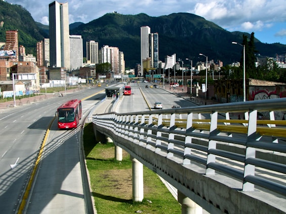 A bustling urban scene featuring a modern cityscape with tall buildings set against a backdrop of lush green mountains. A main roadway shows a dedicated bus lane with several red buses in transit, highlighting efficient public transportation. A pedestrian walkway is visible alongside the road.