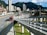 A bustling urban scene featuring a modern cityscape with tall buildings set against a backdrop of lush green mountains. A main roadway shows a dedicated bus lane with several red buses in transit, highlighting efficient public transportation. A pedestrian walkway is visible alongside the road.