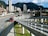 A bustling urban scene featuring a modern cityscape with tall buildings set against a backdrop of lush green mountains. A main roadway shows a dedicated bus lane with several red buses in transit, highlighting efficient public transportation. A pedestrian walkway is visible alongside the road.