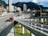 A bustling urban scene featuring a modern cityscape with tall buildings set against a backdrop of lush green mountains. A main roadway shows a dedicated bus lane with several red buses in transit, highlighting efficient public transportation. A pedestrian walkway is visible alongside the road.