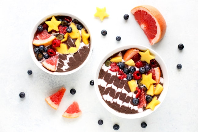 Two smoothie bowls filled with a dark smoothie base, topped with a variety of colorful fruits including star-shaped mango pieces, grapefruit slices, blueberries, raspberries, and other mixed berries. The arrangement is artistic and vibrant, paired with scattered fruit pieces on a white backdrop.