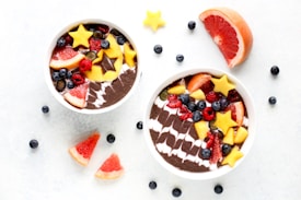 Two smoothie bowls filled with a dark smoothie base, topped with a variety of colorful fruits including star-shaped mango pieces, grapefruit slices, blueberries, raspberries, and other mixed berries. The arrangement is artistic and vibrant, paired with scattered fruit pieces on a white backdrop.