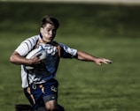 A rugby player wearing a striped jersey and navy shorts is running on a grassy field while holding a rugby ball under his right arm. His expression is focused, and his left arm is extended outward.