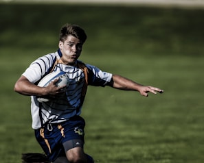 A rugby player wearing a striped jersey and navy shorts is running on a grassy field while holding a rugby ball under his right arm. His expression is focused, and his left arm is extended outward.