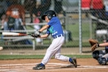 A young baseball player in a blue jersey and white pants is swinging a bat to hit an incoming baseball. The player wears a helmet and gloves, suggesting protective gear necessary for baseball. A catcher is seen behind the batter, kneeling and ready to catch the ball, wearing protective gear including knee pads and a glove. The background features a chain-link fence and blurred spectators.
