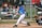 A young baseball player in a blue jersey and white pants is swinging a bat to hit an incoming baseball. The player wears a helmet and gloves, suggesting protective gear necessary for baseball. A catcher is seen behind the batter, kneeling and ready to catch the ball, wearing protective gear including knee pads and a glove. The background features a chain-link fence and blurred spectators.