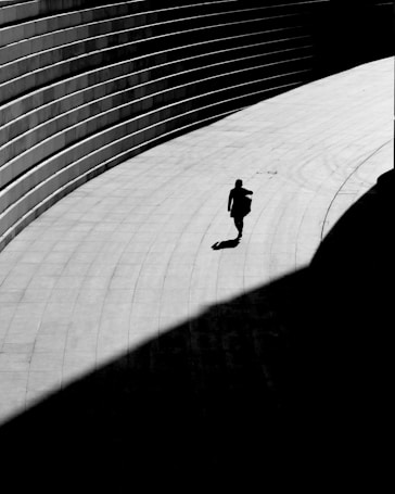A solitary figure walks along a curved, paved pathway, which is partially covered in shadow. The path is adjacent to a series of parallel, horizontal lines formed by steps or seating, creating a dramatic contrast between light and dark areas.