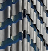 A close-up view of a building facade featuring an intricate pattern of undulating, metallic panels that create a wave-like effect. The design incorporates both reflective and matte surfaces, allowing light to play across the structure, revealing subtle shades of gray and blue.