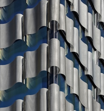 A close-up view of a building facade featuring an intricate pattern of undulating, metallic panels that create a wave-like effect. The design incorporates both reflective and matte surfaces, allowing light to play across the structure, revealing subtle shades of gray and blue.