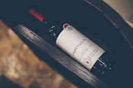 A wine bottle labeled 'CAPÍTULO' with a distinct red cap and a textured white label featuring decorative elements. The bottle lies horizontally inside a dark, wooden wine rack with a blurred background of stone-like texture.