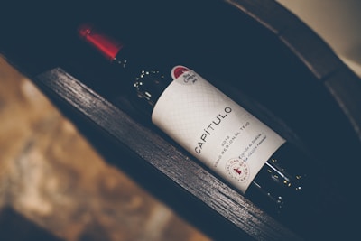 A wine bottle labeled 'CAPÍTULO' with a distinct red cap and a textured white label featuring decorative elements. The bottle lies horizontally inside a dark, wooden wine rack with a blurred background of stone-like texture.