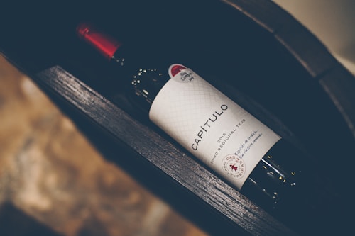 A wine bottle labeled 'CAPÍTULO' with a distinct red cap and a textured white label featuring decorative elements. The bottle lies horizontally inside a dark, wooden wine rack with a blurred background of stone-like texture.