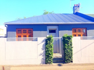 A small, modern house with a flat roof and light-colored exterior walls is surrounded by a simple, minimalist concrete fence. The house features two large wooden-framed windows and a central door. Green climbing plants add a touch of greenery, while the sky appears bright and clear.