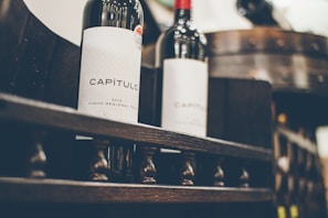 Two wine bottles with the label 'CAPÍTULO 2015 Vinho Regional Tejo' are placed on a wooden rack. The focus is on the intricate design and dark color of the bottles, with a barrel in the blurred background.