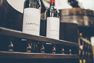 Two wine bottles with the label 'CAPÍTULO 2015 Vinho Regional Tejo' are placed on a wooden rack. The focus is on the intricate design and dark color of the bottles, with a barrel in the blurred background.