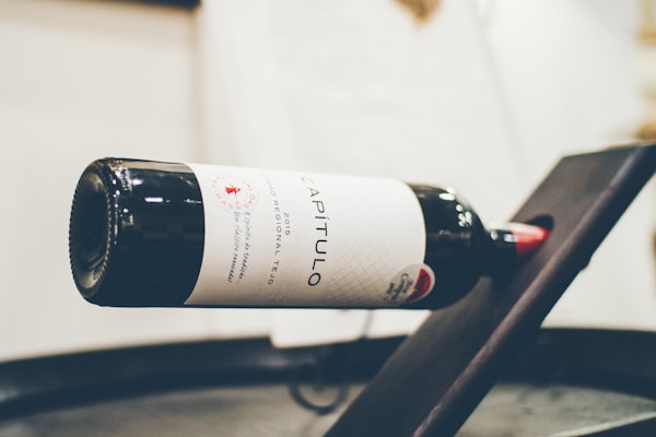 A bottle of wine is displayed horizontally on a rack. The label reads 'Capitulo' and it is a 2015 vintage from the Tejo region. The setting appears to be a wine shop or tasting room with soft lighting and a blurred background.