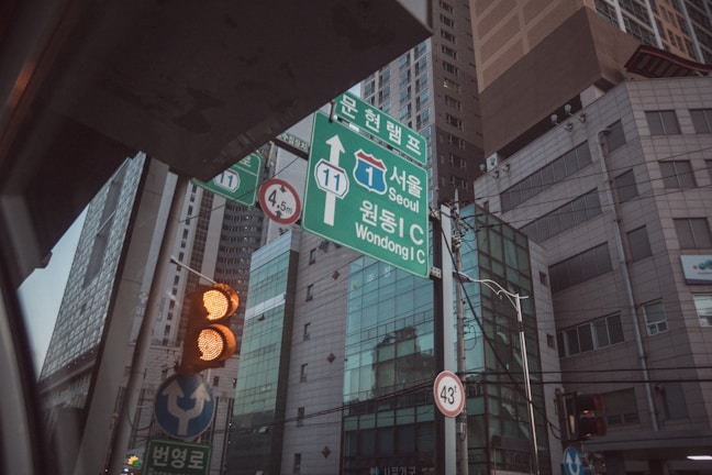 A street scene in an urban area with a focus on road signs and traffic lights. The road signs provide directions to Seoul and Wondong IC, and there are traffic lights showing amber lights. Tall modern buildings and glass windows form the backdrop. Additionally, wires and poles are visible, contributing to the cityscape.