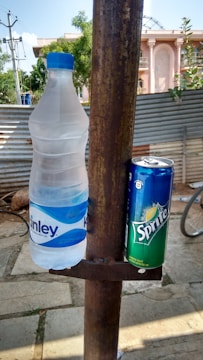 A plastic bottle of Kinley water and a can of Sprite are placed on a rusted metal holder attached to a pole. The background shows an outdoor setting with a corrugated metal fence, greenery, and a partial view of a building.