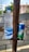 A plastic bottle of Kinley water and a can of Sprite are placed on a rusted metal holder attached to a pole. The background shows an outdoor setting with a corrugated metal fence, greenery, and a partial view of a building.