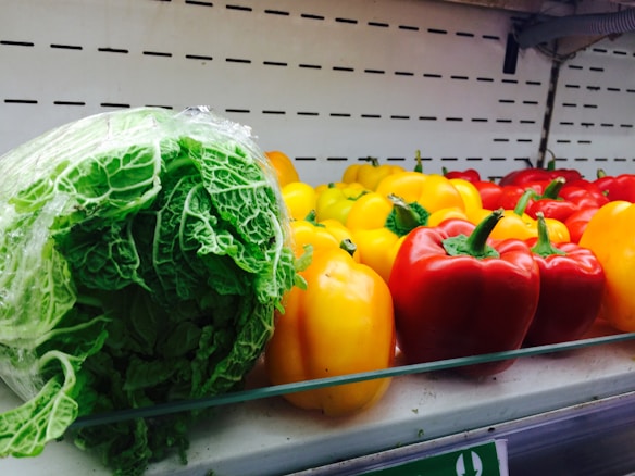 A shelf in a grocery store displays fresh vegetables, including a large, leafy green cabbage wrapped in plastic, and an assortment of vibrant bell peppers in red, yellow, and orange hues.