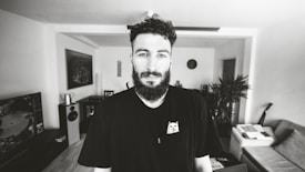 A man with a beard stands in a modern living room. The room features a sofa, indoor plants, a wall clock, and various electronic devices. He wears a black t-shirt with a small cartoon graphic. The setting appears bright and well-organized.