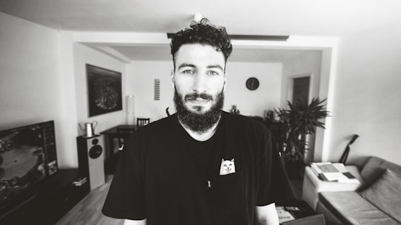A man with a beard stands in a modern living room. The room features a sofa, indoor plants, a wall clock, and various electronic devices. He wears a black t-shirt with a small cartoon graphic. The setting appears bright and well-organized.