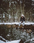 man wearing white knit cap and black jacket sitting on snow covered tree log