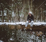 man wearing white knit cap and black jacket sitting on snow covered tree log