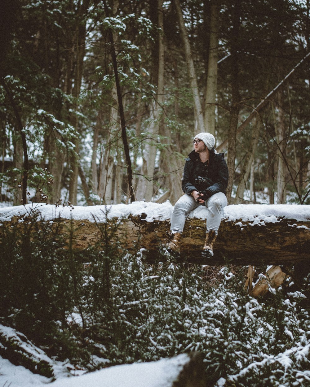 Hombre con gorra de punto blanca y chaqueta negra sentado en un tronco de árbol cubierto de nieve