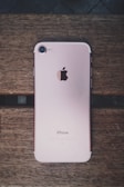 A rose gold smartphone with an Apple logo on the back is placed on a wooden surface. The device is lying flat, showing its camera, flash, and antenna lines.
