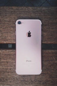 A rose gold smartphone with an Apple logo on the back is placed on a wooden surface. The device is lying flat, showing its camera, flash, and antenna lines.