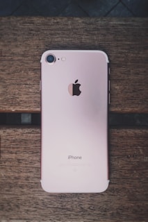 A rose gold smartphone with an Apple logo on the back is placed on a wooden surface. The device is lying flat, showing its camera, flash, and antenna lines.
