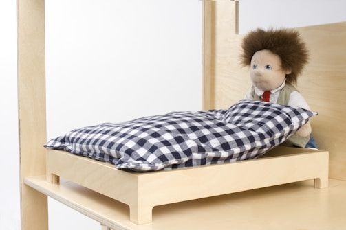 A stuffed doll with a fluffy hairstyle is positioned sitting upright on a small, wooden bed. The bed has a checkered pillow in black and white. The surrounding environment is minimalistic, with light wood tones and a simple design.