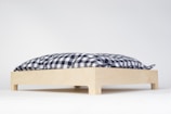 A wooden platform with four small legs supports a blue and white checkered cushion. The cushion appears soft and is positioned on a clean, white background, emphasizing its simplicity and minimalism.