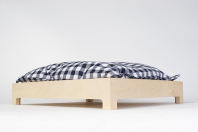 A wooden platform with four small legs supports a blue and white checkered cushion. The cushion appears soft and is positioned on a clean, white background, emphasizing its simplicity and minimalism.