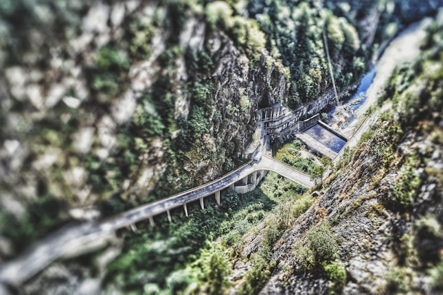A scenic view of a winding road traversing through a steep, rocky gorge with lush greenery. In the background, a river flows alongside the road, and a bridge structure is visible.