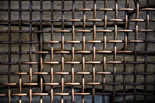 A close-up view of a rusty metal grid pattern, with a weathered stone wall behind it. The grid is composed of interwoven metal rods forming a lattice structure. The lighting casts shadows, emphasizing the texture of both the metal and the stone.