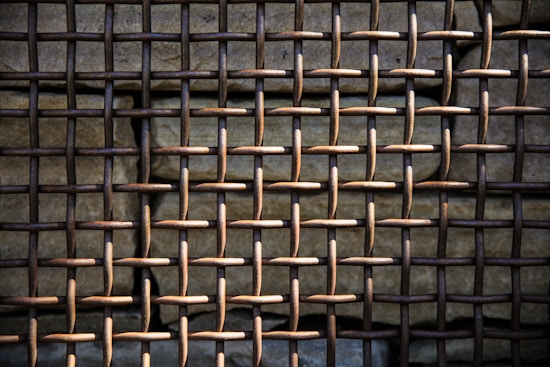 A close-up view of a rusty metal grid pattern, with a weathered stone wall behind it. The grid is composed of interwoven metal rods forming a lattice structure. The lighting casts shadows, emphasizing the texture of both the metal and the stone.