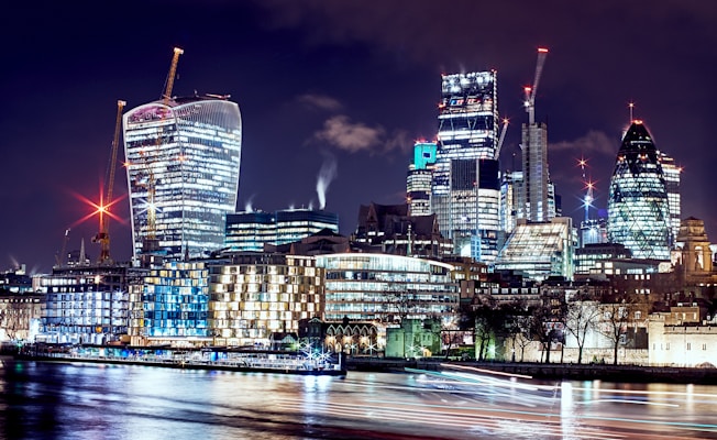 A night-time cityscape showcasing a diverse array of modern skyscrapers illuminated against the night sky. The buildings are distinct in shape and size, with lights glowing from their interiors and construction cranes visible. The reflective surface of a river in the foreground captures the vibrant lights.