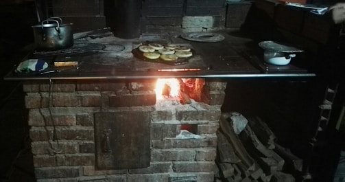 A rustic brick stove with a metal top is active, with a fire burning in the lower compartment. Cooking utensils, including a pot and a baking sheet with food, are scattered across the surface.