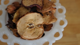 A decorative bowl crafted from white ceramic with circular cutouts is filled with a variety of dried fruit slices, including apple and potentially other dried botanicals.