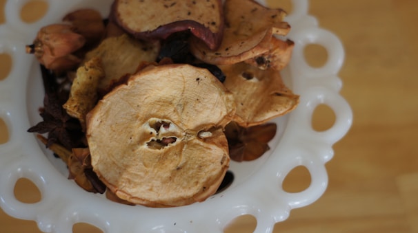 A decorative bowl crafted from white ceramic with circular cutouts is filled with a variety of dried fruit slices, including apple and potentially other dried botanicals.