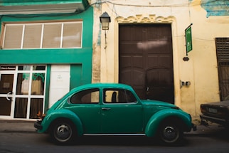 A classic green Volkswagen Beetle is parked along a street in front of a building with green and beige walls. The building features a large dark brown door and modern window design. An old street lamp hangs above the car, and there are other parked vehicles partially visible.